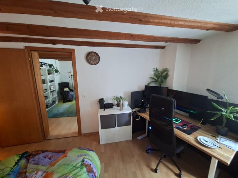 A furnished room with a desk setup, including a chair, desk, multiple screens, keyboard, mouse, and a small cabinet. There is a colorful bedspread visible, a clock on the wall, and a plant on the desk.