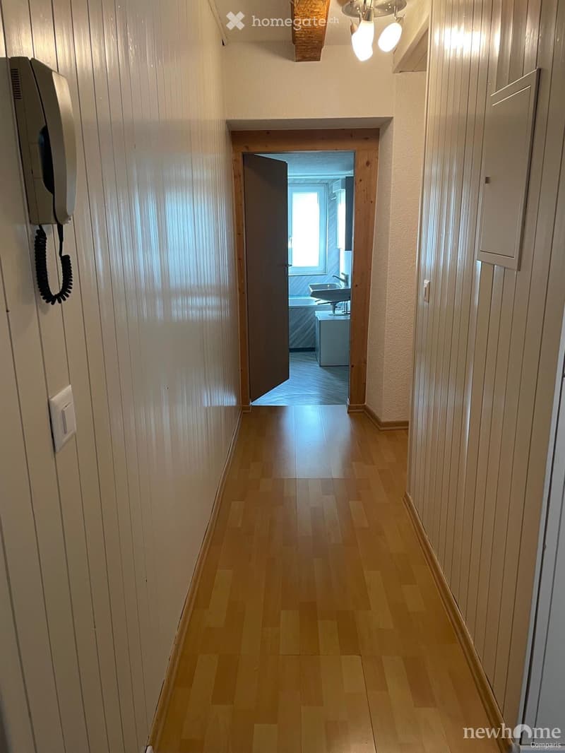 A narrow hallway with wooden flooring and light-colored walls, featuring a telephone on the left and a partially open door leading to a bathroom with a sink and bathtub visible.