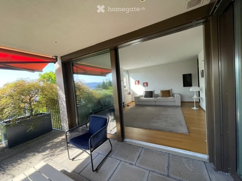 Balcony with a view, featuring a single chair and a large sliding glass door leading to a living room with a sofa and a lamp.