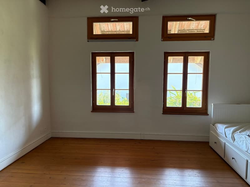 Empty bedroom with wooden flooring, white walls, and four windows with wooden frames. A single bed with white bedding is positioned against the wall.