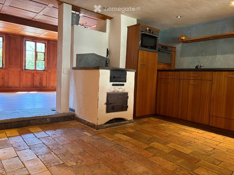 Kitchen with wooden cabinets, a built-in oven, and a stone stove. The floor is made of terracotta tiles, and there is a large window to the left.