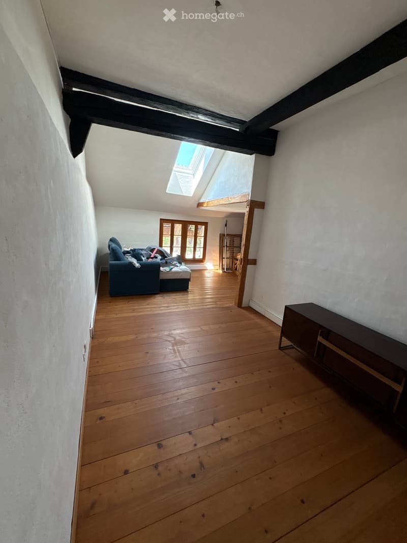 A furnished room with wooden flooring, white walls, and exposed wooden beams. Features include a sofa, a small table, and a chair, with a window and a skylight providing natural light.