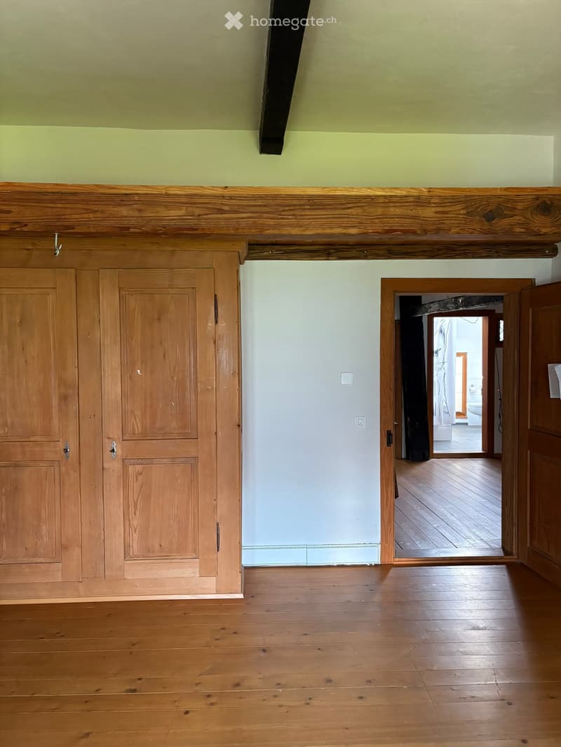 Empty room with wooden flooring, light green walls, and a wooden cabinet. An open doorway leads to another room with visible wooden flooring and a glimpse of a bathroom.