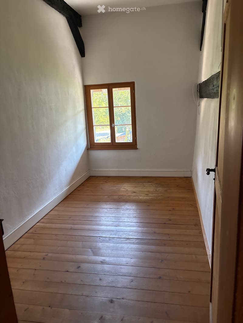 Empty room with wooden flooring and white walls, featuring a small window with wooden frame and a door visible on the right side.