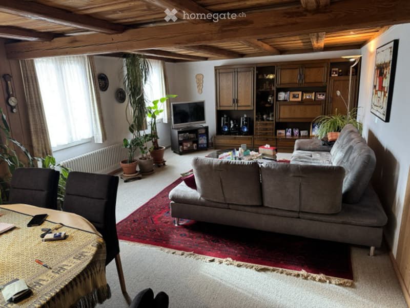 Cozy living room with exposed wooden beams, large window, grey sectional sofa, coffee table, TV stand, dining table with chairs, potted plants, and wooden cabinetry.