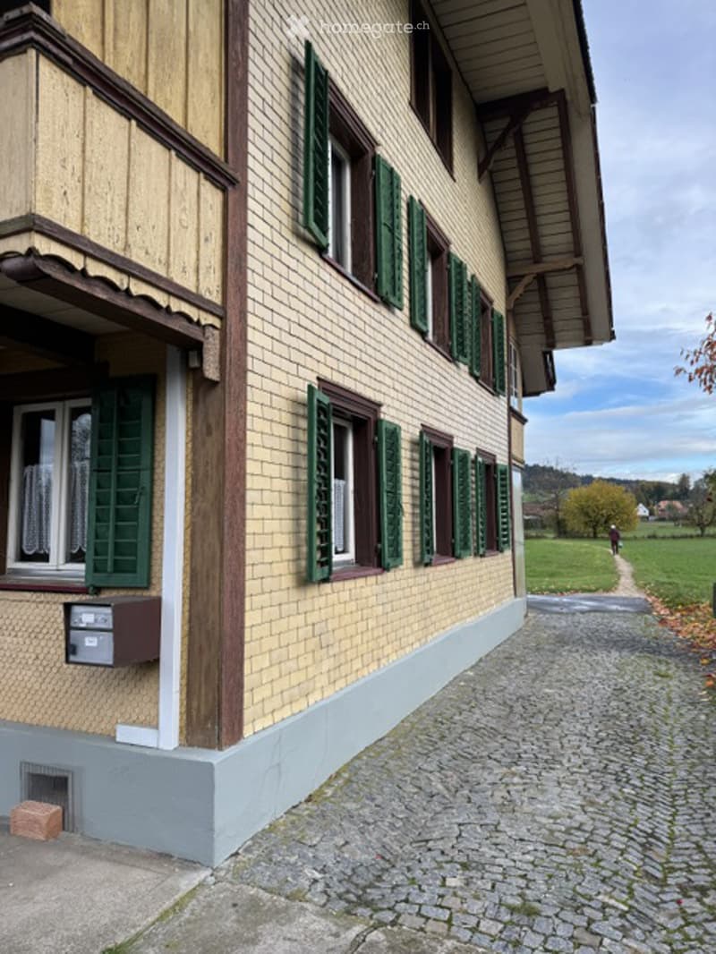 A two-story building with a mix of wooden and brick facades, green shutters, and a small balcony on the first floor. The building is situated on a cobblestone pathway with a grassy area and trees in the background.