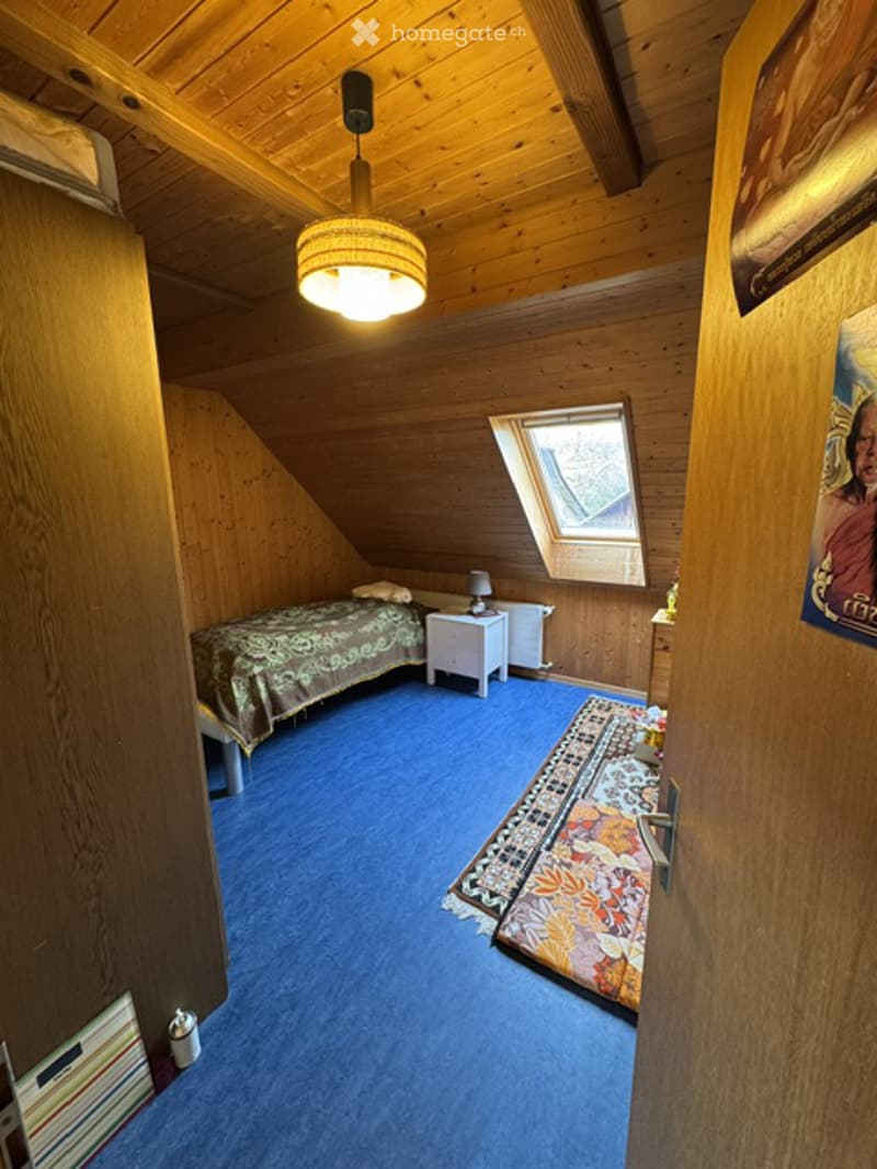 Cozy attic bedroom with wooden walls and ceiling, blue carpet, a bed with patterned bedding, a nightstand with a lamp, and a colorful rug on the floor.