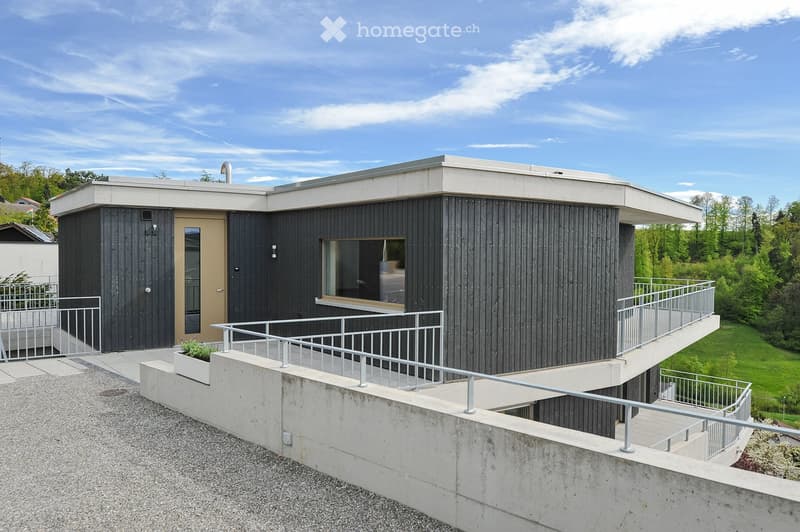 Modern, single-story building with dark wooden siding and white concrete accents, featuring a raised ground floor, a ramp, and a small balcony with railings.