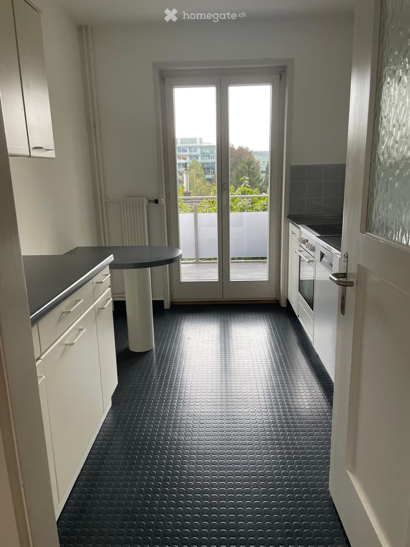 Modern kitchen with white cabinets, a black countertop, a round table, and a stove. The floor has a textured black pattern, and there is a large glass door leading to a balcony.