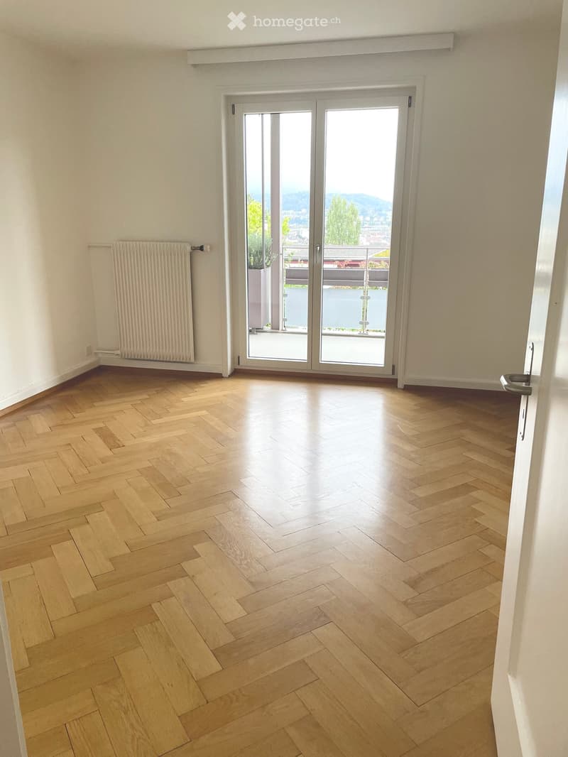 Empty room with light wooden herringbone flooring, white walls, and a large sliding glass door leading to a balcony with a view.