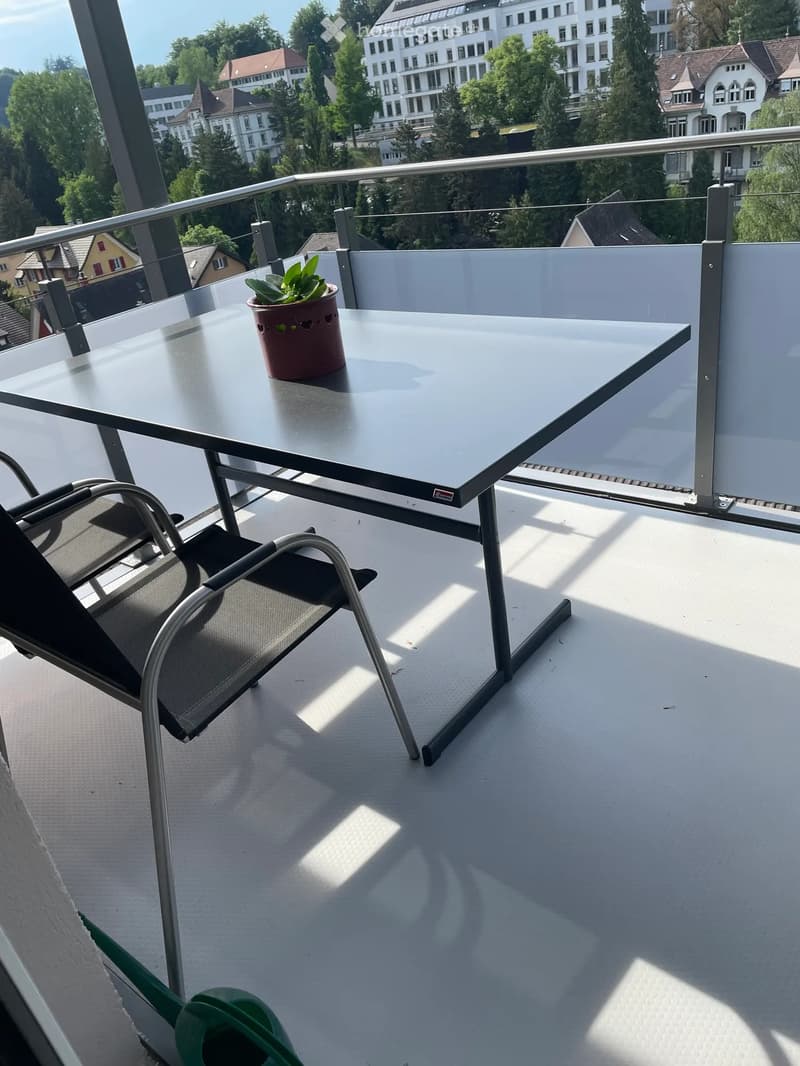 Balcony with a glass table, a chair, and a small potted plant, overlooking a cityscape with buildings and trees.