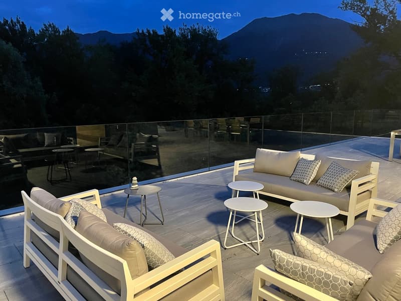 Modern outdoor terrace with white lounge furniture, small tables, and a scenic mountain view at dusk.