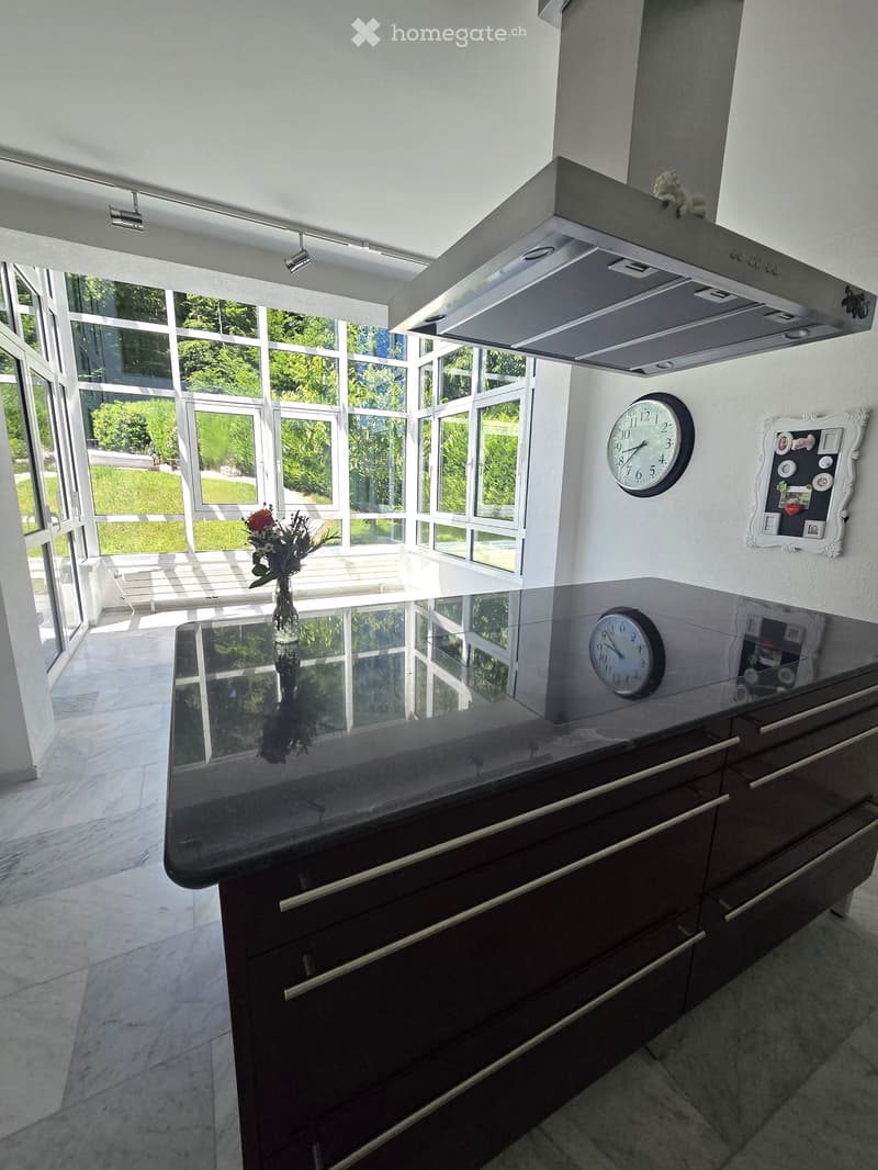 Modern kitchen with dark wooden cabinets, a black countertop, a vase with flowers, a wall clock, and a picture frame on the wall. Large windows provide natural light.