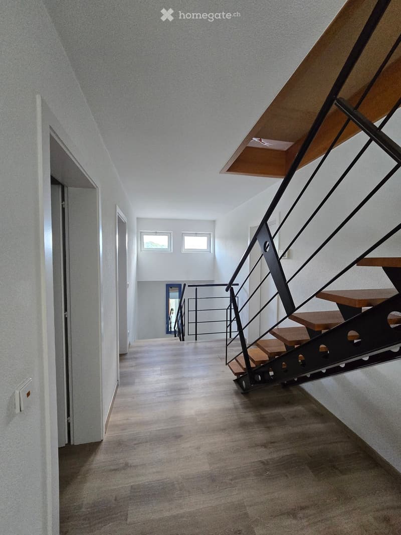 Modern hallway with wooden flooring, white walls, and a staircase with a black metal railing leading upwards. Two windows provide natural light.