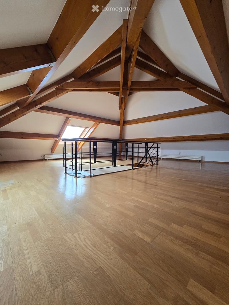 Empty attic space with wooden beams, wooden floor, and a small metal staircase leading to a loft area.
