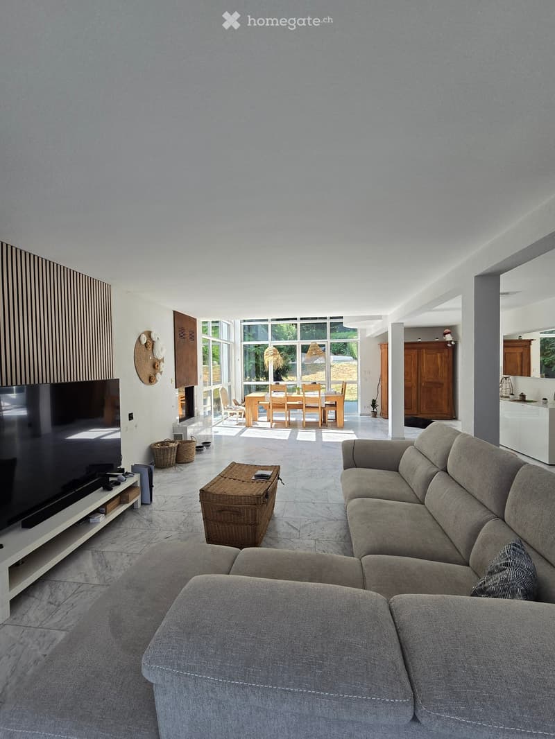 Bright living room with large windows, grey sectional sofa, coffee table, TV, and dining area visible through an open space.