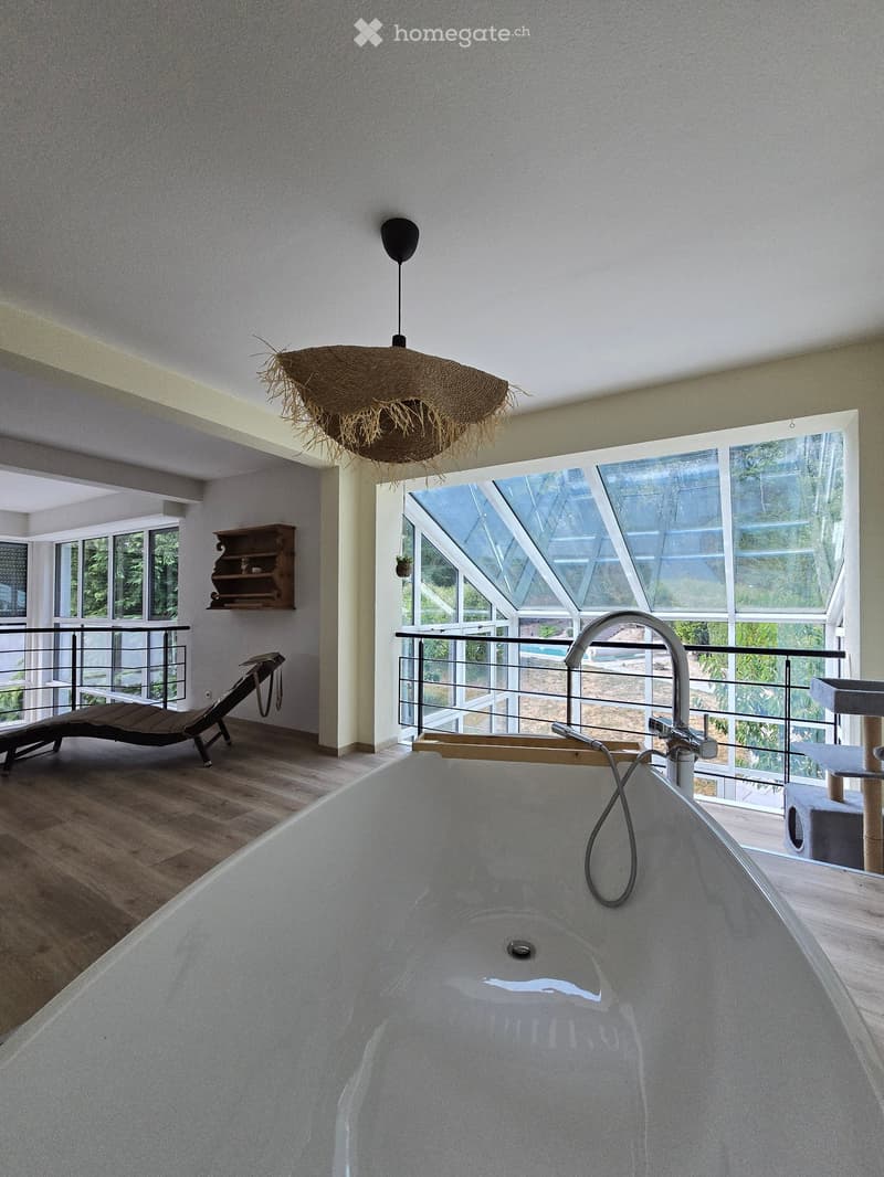 Modern bathroom with a large freestanding bathtub, sleek faucet, cat tree, wooden shelf, lounge chair, and a woven pendant light fixture.
