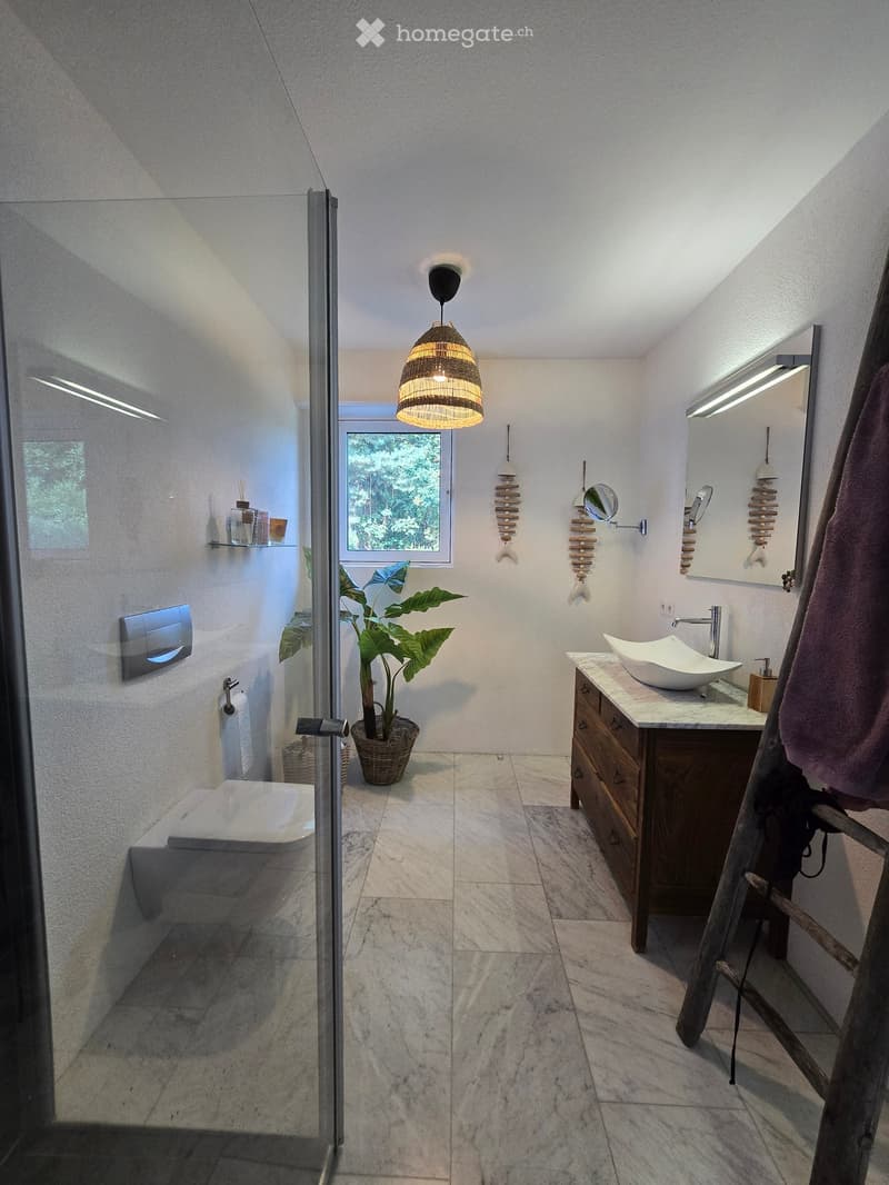 Modern bathroom with a sleek sink, bathtub, and large window. Features a hanging wicker light fixture, decorative wall hangings, and a potted plant in a wicker basket.