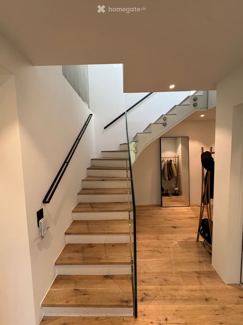 Modern hallway with wooden flooring, staircase with glass railing, coat rack, and mirror reflecting coats and shoes.