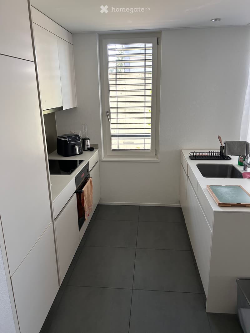 Modern kitchen with white cabinets, dark grey tiles, and stainless steel appliances. Features a coffee maker, oven, sink, dish rack, and cutting board.