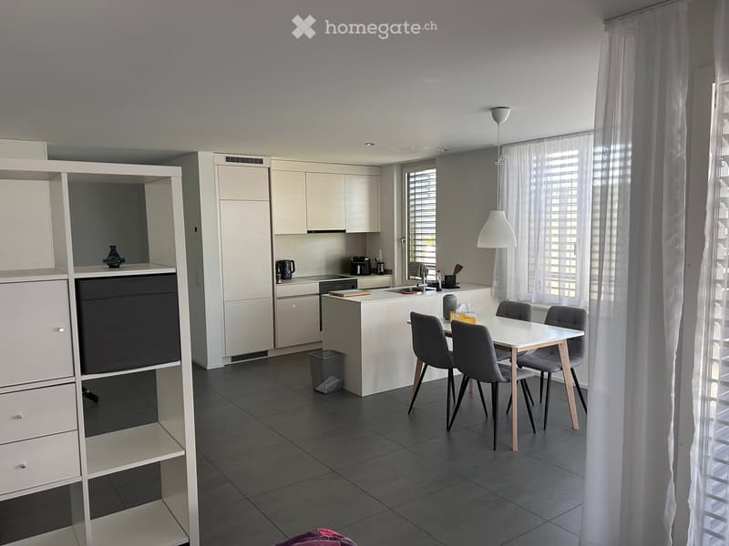 Modern kitchen with white cabinets, a central island, and a dining area with a table and chairs. The space features a sink, coffee maker, and a hanging lamp.