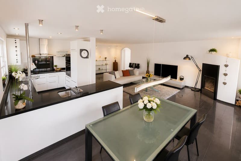 Open-plan living and dining area with a modern kitchen, white cabinets, black countertops, a glass dining table with chairs, a white sofa, a fireplace, and decorative vases with flowers.