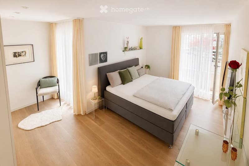 Modern bedroom with a large bed, nightstand, chair, and decorative items including a vase with a rose and a framed picture on the wall.