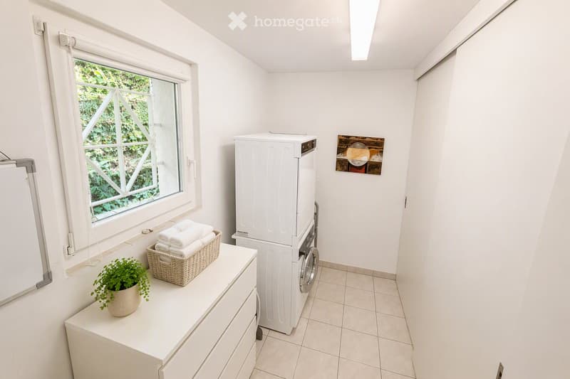 Bright bathroom with white walls, tiled floor, and a window. Features a stacked washing machine and dryer, a wicker basket with towels, a small potted plant on a cabinet, and a framed picture on the wall.