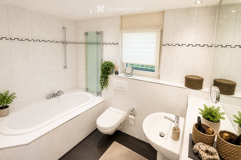 Modern bathroom with white tiles, bathtub, toilet, sink, and decorative plants. Features a large window with blinds and wicker baskets on the windowsill.