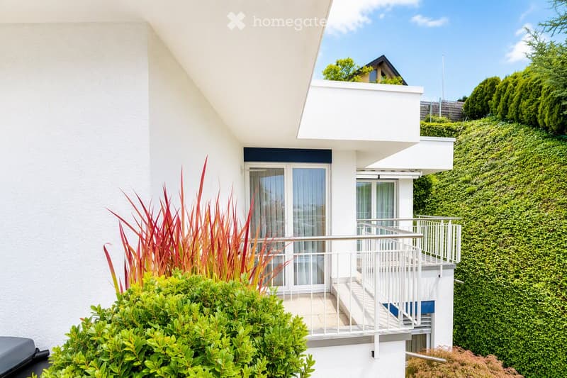 Balcony with white railing, glass doors leading inside, surrounded by greenery and shrubs.
