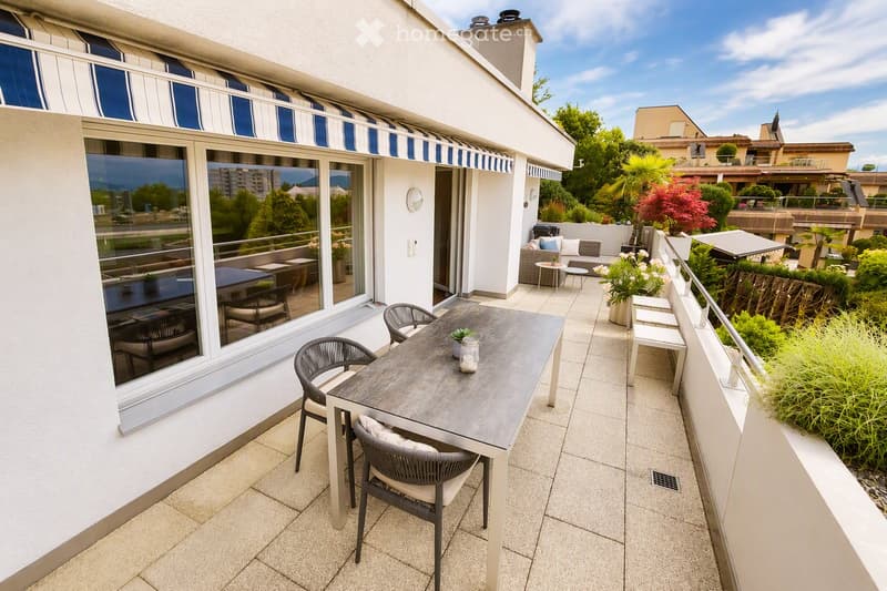 Balcony with a dining table and chairs, overlooking a landscaped area with greenery and other buildings.