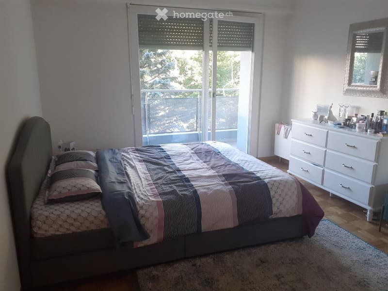 Cozy bedroom with a large bed, patterned bedding, a dresser with various items, a mirror, and a small table with a chair. The room features wooden flooring and a view through a sliding glass door.