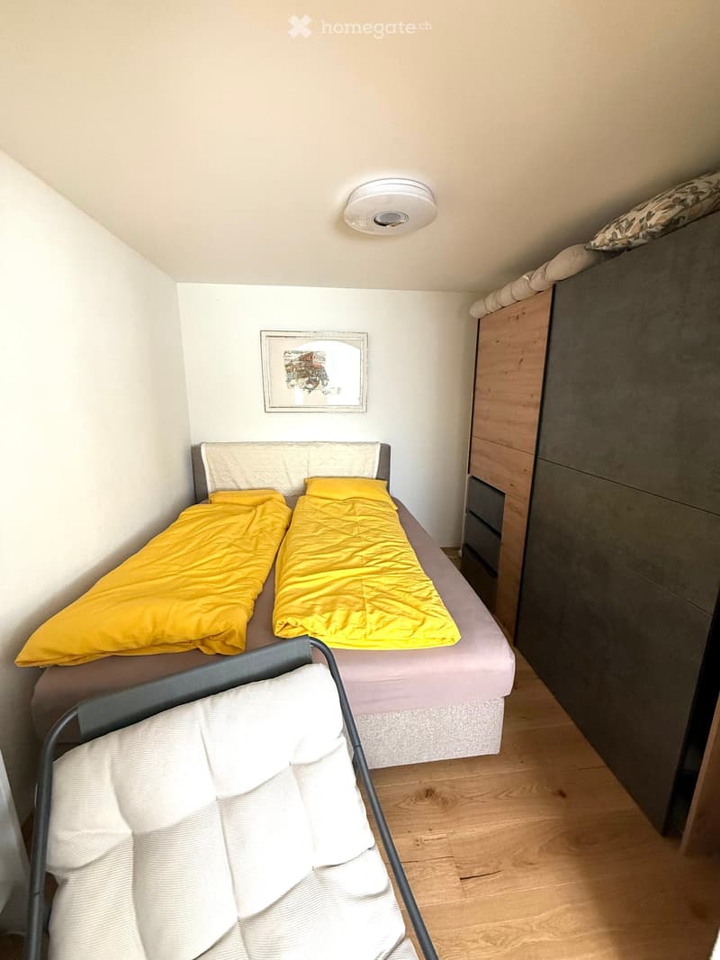 Cozy bedroom with a double bed, yellow blankets, a wooden wardrobe, and a chair with a white cushion.