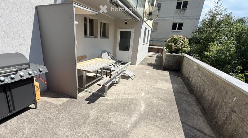 Balcony with a wooden table, bench, chairs, and a grill, surrounded by greenery and a building entrance.