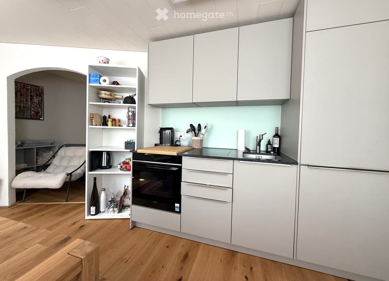 Modern kitchen with white cabinets, a built-in oven, a sink with a faucet, and a small shelf unit holding various items. A white chair and a small wooden stool are also visible.