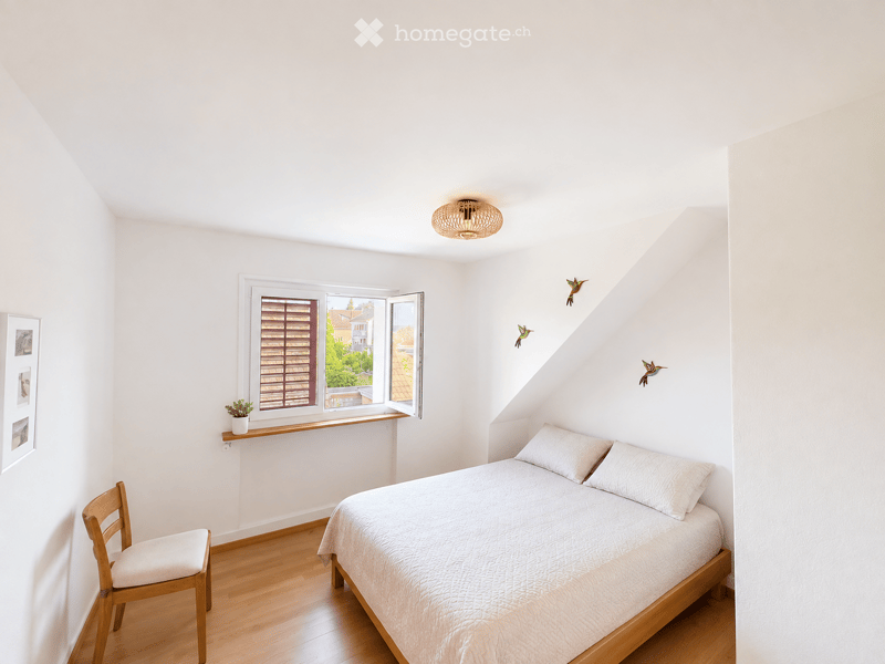 Bright, minimalist bedroom with wooden flooring, a single bed, a wooden chair, a small potted plant on a windowsill, and decorative wall art.