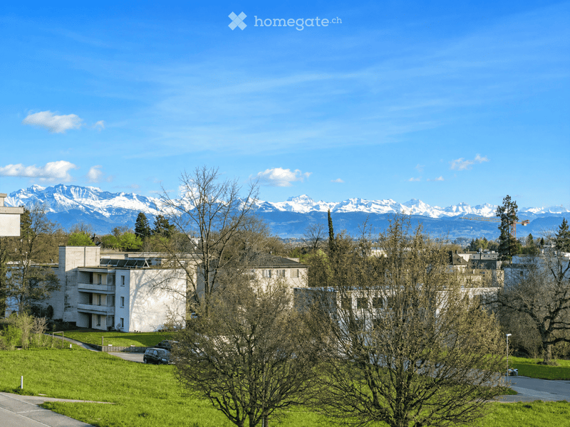 Modern multi-story residential building with white walls and balconies, surrounded by green lawn and trees, with snow-capped mountains in the background.
