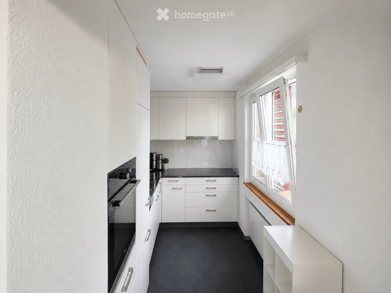 Modern kitchen with white cabinets, black countertop, and stainless steel appliances. Features an oven, sink, and built-in storage. Natural light from a window with a wooden sill.