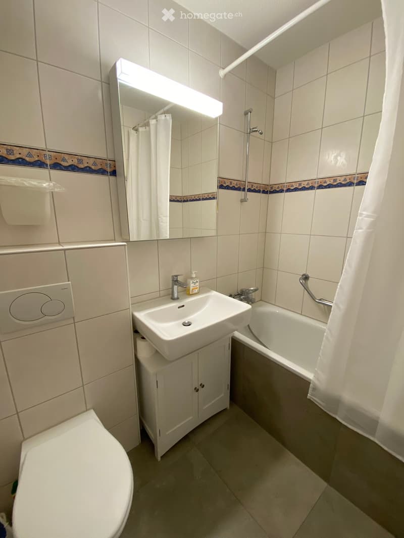 Bathroom with white tiles, a sink with a mirror above it, a toilet, a bathtub with a shower curtain, and a small cabinet under the sink.