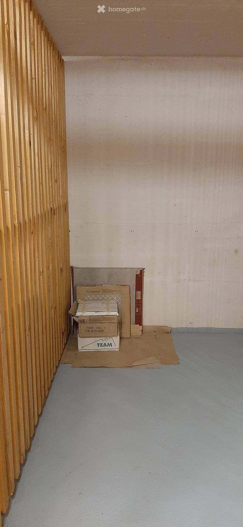 Empty room with bare walls and a grey floor, featuring a wooden panel on one side and a few cardboard boxes stacked in the corner.