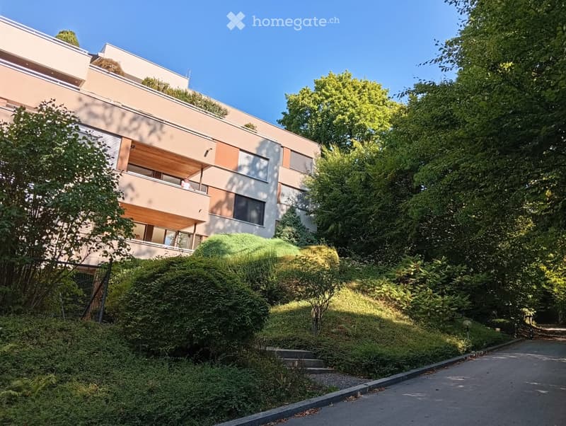 Modern multi-story building with balconies and large windows, surrounded by lush greenery and trees.