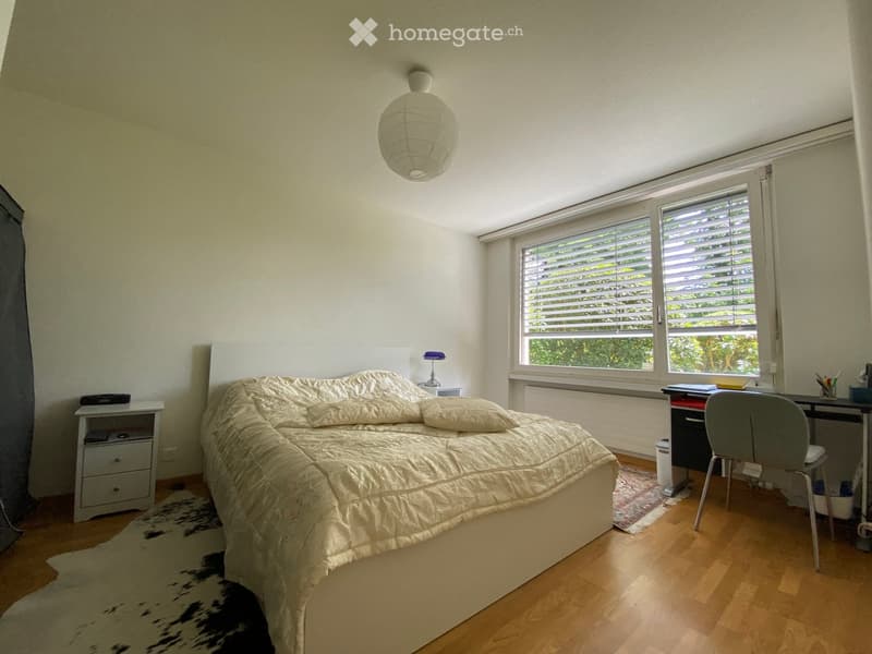 Bright bedroom with wooden flooring, large window with blinds, bed with beige bedding, nightstand, desk with chair, and a hanging paper lantern light fixture.