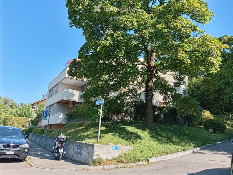 Modern multi-story building with a raised ground floor, surrounded by greenery and trees. A parked car and motorcycle are visible on the street.