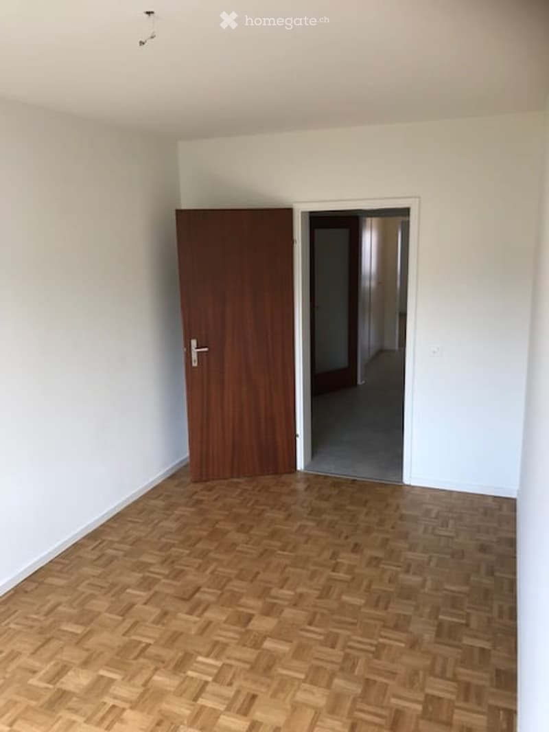 Empty room with wooden herringbone flooring, white walls, and a closed wooden door leading to another space.