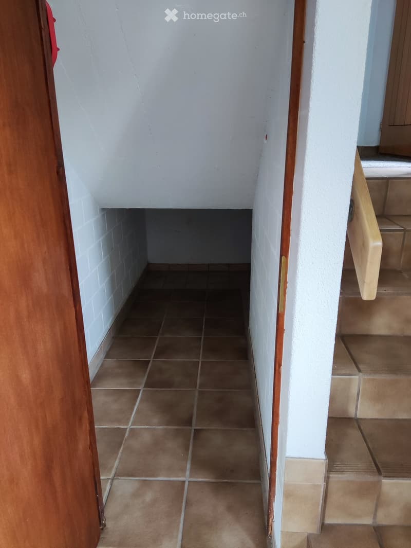A narrow hallway with tiled floor, white walls, and a wooden door on the left. A staircase with wooden handrails is visible on the right.