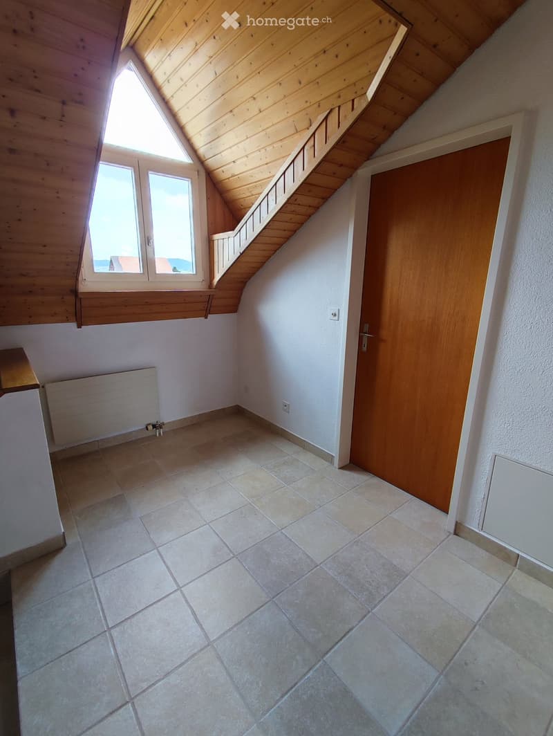 Empty room with tiled floor, white walls, wooden door, and a window with a view. The ceiling is slanted with wooden beams.