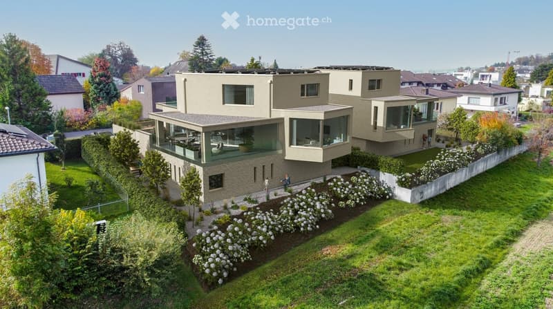 Modern two-story house with large windows, flat roof, and surrounding garden with neatly trimmed lawn and flower beds.