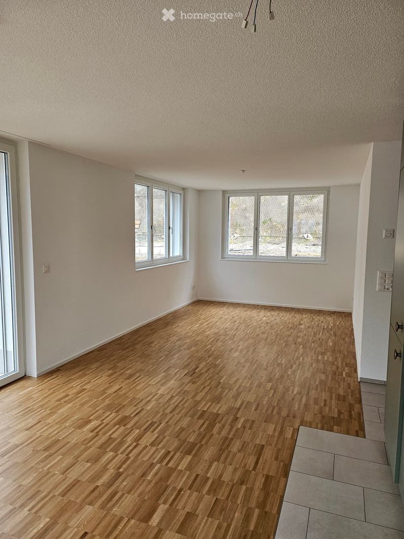 Empty room with wooden flooring, white walls, and large windows, featuring a door and electrical outlets.