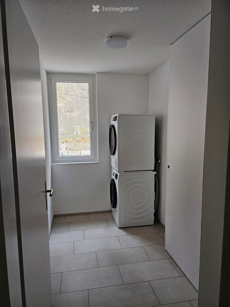 A small bathroom with tiled floors and white walls, featuring a stacked washing machine and dryer unit near a frosted window.