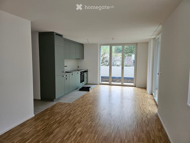 Empty kitchen with light grey cabinets, a sink, and a sliding glass door leading to an outdoor area. The floor is a combination of wooden and tiled surfaces.